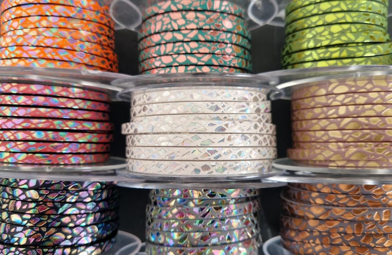 May include: Nine spools of flat leather cord arranged in a grid, each featuring a distinct metallic or iridescent cracked pattern. The colors include orange, teal, lime green, pink, white, gold, black, silver, and bronze, all wound on clear plastic.