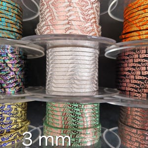 Könnte beinhalten: Spulen mit dekorativem Band in verschiedenen Farben und Mustern, darunter Orange, Rosa, Schwarz und Grün. Das Band ist 3 mm breit und wird auf transparenten Kunststoffspulen präsentiert.