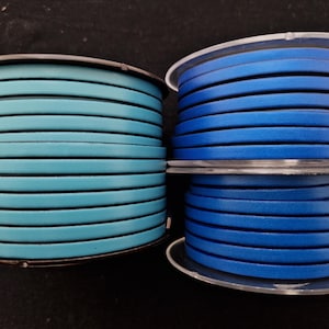 May include: Two spools of leather cord in shades of blue. One spool is a light teal blue, and the other two are a darker, royal blue. The cord is flat and appears to be approximately 3 mm wide. The spools are on a dark surface.