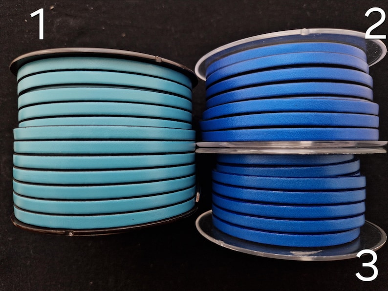 May include: Two spools of leather cord in shades of blue. One spool is a light teal blue, and the other two are a darker, royal blue. The cord is flat and appears to be approximately 3 mm wide. The spools are on a dark surface.