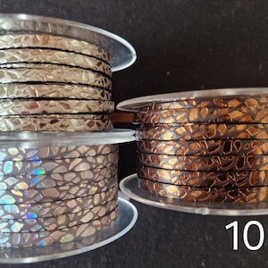 May include: Three spools of flat cord with a textured, cracked metallic finish. Spool 8 features a silver tone, spool 9 displays an iridescent holographic pattern, and spool 10 shows a deep copper color. Each spool is labeled with a white number.