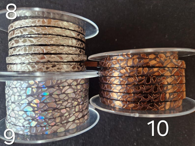 May include: Three spools of flat cord with a textured, cracked metallic finish. Spool 8 features a silver tone, spool 9 displays an iridescent holographic pattern, and spool 10 shows a deep copper color. Each spool is labeled with a white number.