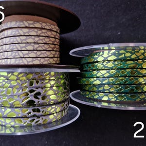 May include: Three spools of flat leather cord labeled 26, 27, and 28. Spool 26 features a beige and brown cracked pattern. Spool 27 displays a metallic lime green and grey cracked design. Spool 28 shows a metallic emerald green and gold cracked pattern.