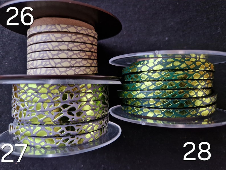 May include: Three spools of flat leather cord labeled 26, 27, and 28. Spool 26 features a beige and brown cracked pattern. Spool 27 displays a metallic lime green and grey cracked design. Spool 28 shows a metallic emerald green and gold cracked pattern.