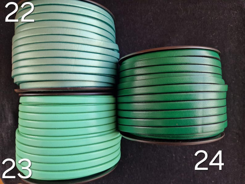 May include: Four spools of green leather cord. The spools are labelled with the numbers 22, 23, and 24. The cord is flat and tightly wound. The background is a dark colour. The cord is suitable for crafts.