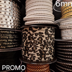 May include: A collection of spools featuring various ribbons and trims, including silver star-patterned, white studded, gold houndstooth, black with 'Have a lovely day' text, leopard print, and black studded designs, with a 6mm label visible.