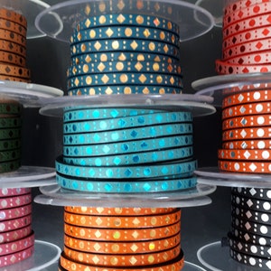 May include: Spools of decorative trim with a variety of colours and patterns. The trim features a repeating pattern of circles and squares with a metallic finish.