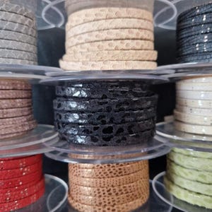 Peut inclure: Bobines de cordon en cuir de différentes couleurs, dont gris, beige, noir et rouge. Les cordons ont une surface texturée et à motifs, et sont enroulés sur des bobines en plastique transparent. L'image présente des fournitures artisanales.