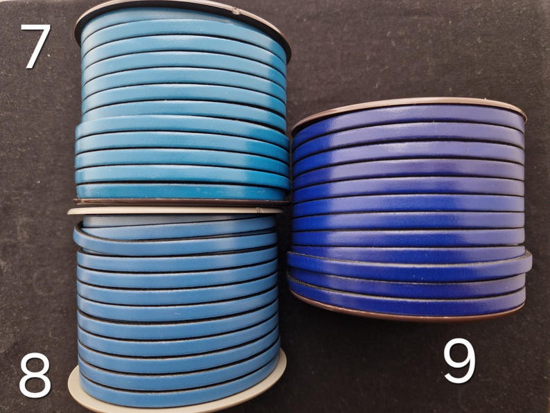 May include: Three spools of flat leather cord in shades of blue. The spools are stacked, with the top one in a lighter blue, the middle in a medium blue, and the bottom in a darker blue. The cords are neatly wound.