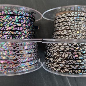May include: Four spools of flat leather cord labeled 1 through 4. Spool 1 is black with iridescent shards. Spool 2 is light gray with iridescent shards. Spool 3 is light gray with silver shards. Spool 4 is black with silver and iridescent shards.