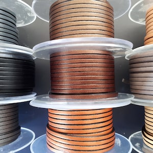 Puede incluir: Múltiples carretes de cordón de cuero plano dispuestos en una cuadrícula. Los carretes presentan varios colores, incluyendo negro, marrón oscuro, marrón medio, bronceado y gris. Cada carrete es de plástico transparente y contiene cordón.