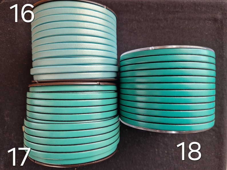 May include: Three spools of teal and turquoise leather cord. The flat cord is approximately 3mm wide. The spools are labelled with the numbers 16, 17, and 18. The cord is wound neatly on the spools.