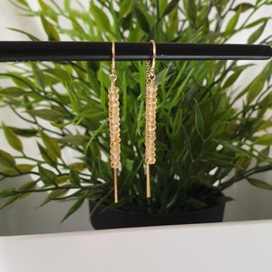 Puede incluir: Un par de pendientes de oro con una cadena larga y fina de pequeñas cuentas de color amarillo claro. Los pendientes cuelgan de una barra negra.