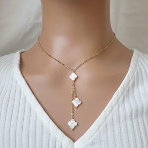 May include: A gold-tone necklace featuring a fine chain and three white, clover-shaped pendants. The necklace is shown on a person wearing a white V-neck top. The pendants have a pearlescent finish, creating a subtle shimmer.
