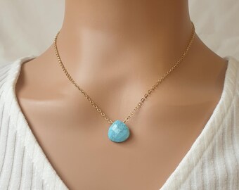 May include: A gold-colored chain necklace with a single, teardrop-shaped, light blue gemstone pendant. The necklace is worn on a person wearing a white ribbed shirt.