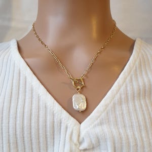 Op de afbeelding: Goudkleurige ketting met een grote, onregelmatig gevormde parel hanger. De ketting heeft een toggle sluiting en een ketting met rechthoekige schakels. De hanger heeft een crèmewitte kleur en een subtiele glans. De ketting wordt gedragen door een persoon die een witte geribbelde top draagt.