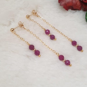 May include: A pair of gold earrings with a dangling chain and three purple gemstone beads.