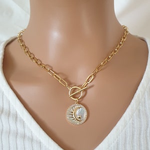 May include: A gold chain necklace with a toggle clasp and a round pendant featuring a sun and moon design with a white shell inlay and clear crystals.