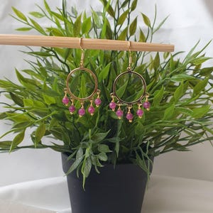 Op de afbeelding: Goudkleurige oorringen met bungelende roze kralen. De oorbellen worden gepresenteerd tegen een achtergrond van groen gebladerte in een zwarte pot en een witte doek. Eenvoudig, elegant ontwerp.