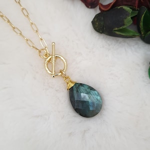 Könnte beinhalten: Goldfarbene Halskette mit einem tropfenförmigen, irisierenden blau-grünen Edelsteinanhänger. Der Anhänger ist mit Draht umwickelt und hängt an einem Knebelverschluss. Die Kette hat große, ovale Glieder.