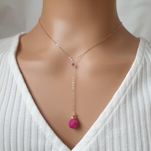 May include: A gold-colored necklace with a Y-shaped design. The necklace features a small, round, pink gemstone at the top and a larger, faceted, pink gemstone pendant at the bottom. The necklace is worn on a person wearing a white shirt.