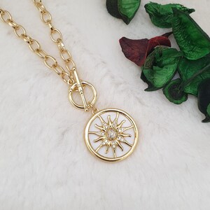 May include: Gold-coloured necklace with a circular pendant featuring a sunburst design. The pendant has a white background with a sun design and a central cluster of small, sparkling stones. The chain is a chunky link style.