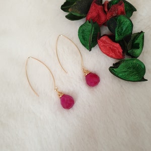 Puede incluir: Un par de pendientes de oro con gotas de piedras preciosas rosas. Los pendientes están sobre una superficie blanca y peluda.