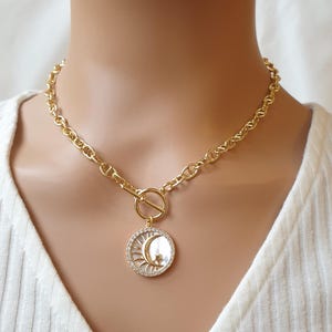 Puede incluir: Un collar de cadena dorada con cierre de palanca y un colgante redondo con un diseño de luna creciente y estrella con brillantes transparentes.