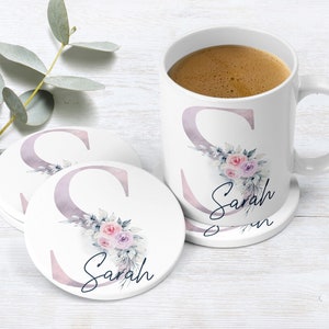 Peut inclure: Une tasse en céramique blanche avec un motif floral et le nom "Sarah" imprimé dessus. La tasse est posée sur un dessous-de-verre assorti avec le même motif. Deux autres dessous-de-verre sont visibles en arrière-plan.