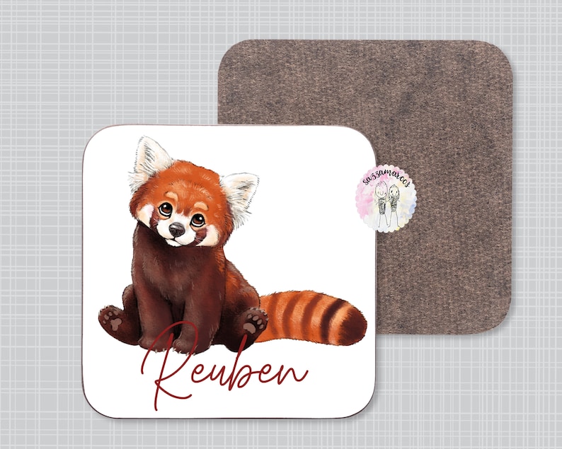 Puede incluir: Un posavasos blanco con una ilustraci&oacute;n de un panda rojo y el nombre "Reuben" escrito en cursiva roja. El posavasos tiene un respaldo de corcho marr&oacute;n.