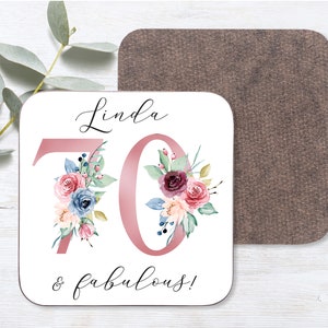 May include: White coaster with a pink number 70 decorated with watercolor flowers. The text "Linda & fabulous!" is written below the number.