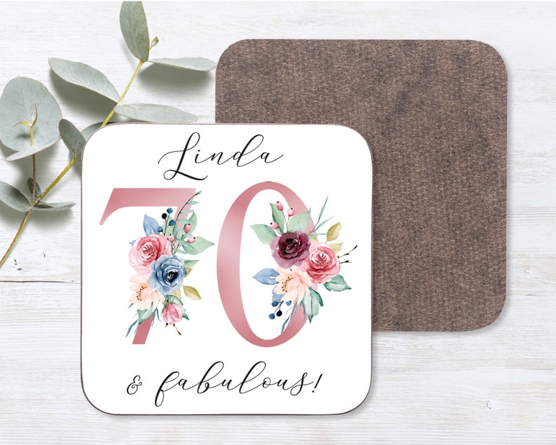 May include: White coaster with a pink number 70 decorated with watercolor flowers. The text "Linda & fabulous!" is written below the number.