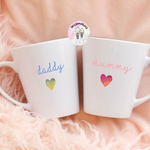 May include: Two white ceramic mugs with handles. The mug on the left says "daddy" in blue script with a yellow and green heart. The mug on the right says "mummy" in pink script with a pink and red heart.