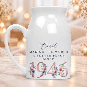 May include: A white ceramic mug with a handle featuring the name Carol in dark blue script, followed by the text MAKING THE WORLD A BETTER PLACE SINCE, and the year 1946 in large rose gold numbers decorated with blue and pink floral accents.