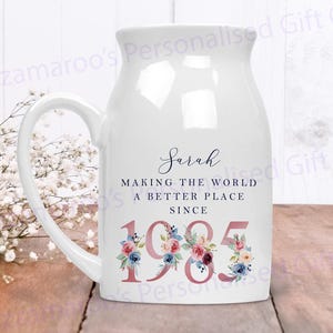 Op de afbeelding: Witte keramische mok met een handvat, versierd met een bloemenontwerp en de tekst "Sarah Making the World a Better Place Since 1985".