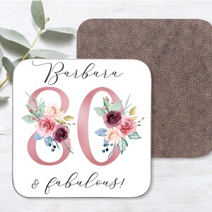May include: White square coaster with the name "Barbara" and the number "80" in pink with floral designs. The text "& fabulous!" is written below the number. The coaster has a brown cork backing.