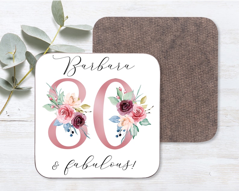 May include: White square coaster with the name "Barbara" and the number "80" in pink with floral designs. The text "& fabulous!" is written below the number. The coaster has a brown cork backing.
