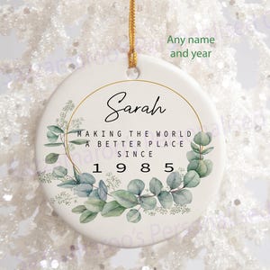 May include: White ceramic bauble with a gold circle and green eucalyptus leaves. The text reads "Sarah Making the World a Better Place Since 1985".