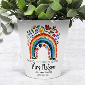 Op de afbeelding: Witte keramische plantenpot met een regenboogontwerp en kleurrijke bloemen. De pot heeft de tekst "THANK YOU FOR HELPING ME TO GROW Mrs Nelson Love From Amelie x 2024" erop gedrukt.