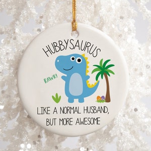 Peut inclure: Ornement de Noël rond en céramique blanche avec une boucle dorée. Il présente une illustration de dinosaure bleu et le texte "Hubbysaurus" et "Like a normal husband, but more awesome."