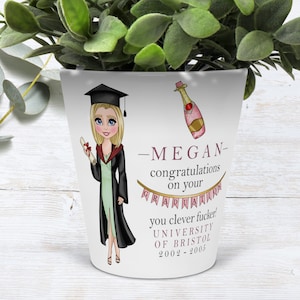 May include: White ceramic planter with a pink and gold illustration of a graduate wearing a cap and gown, holding a diploma, and a bottle of champagne. The text reads "Megan - congratulations on your graduation you clever fucker! University of Bristol 2002 - 2005".