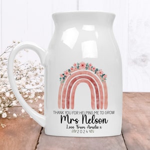 Op de afbeelding: Witte keramische melkkan-mok met een roze en oranje aquarel regenboog met roze bloemen. De mok heeft een handvat en zegt "Thank you for helping me to grow Mrs. Nelson Love from Amelie x 2024"