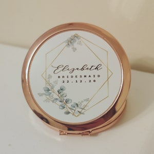 Puede incluir: Un espejo compacto de oro rosa con fondo blanco y un diseño geométrico. El diseño presenta un contorno dorado de una forma de diamante con hojas de eucalipto y el texto "Elizabeth Bridesmaid 22.12.20".