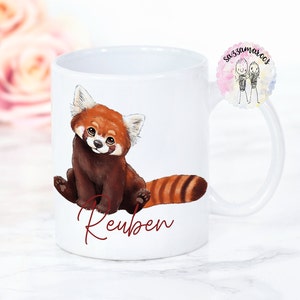 Puede incluir: Taza de cer&aacute;mica blanca con una ilustraci&oacute;n de un panda rojo y el nombre "Reuben" escrito en cursiva roja.