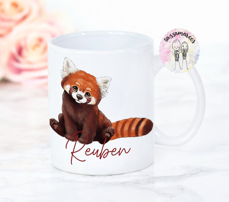 Puede incluir: Taza de cer&aacute;mica blanca con una ilustraci&oacute;n de un panda rojo y el nombre "Reuben" escrito en cursiva roja.