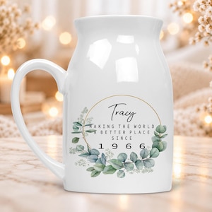 Peut inclure: Un mug en céramique blanche avec un bord évasé et une anse. L'avant présente un cadre circulaire doré entourant le texte Tracy, MAKING THE WORLD A BETTER PLACE SINCE 1966, agrémenté d'une illustration aquarelle de feuilles d'eucalyptus vertes.