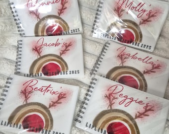 May include: Six spiral-bound notebooks with white covers, each featuring a personalized name in red script, a graphic of a reindeer with a red nose, and the printed text "LAPLAND ADVENTURE 2025" at the bottom of each individual notebook cover.