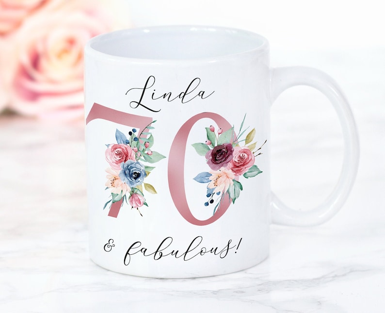 Puede incluir: Taza de cer&aacute;mica blanca con un dise&ntilde;o floral rosa y el texto "Linda 70 & fabulosa!"