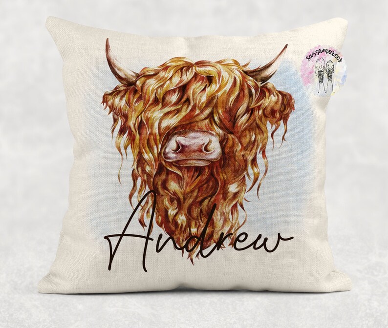 May include: A white pillow with a watercolor illustration of a Highland cow with long, brown hair. The cow's face is visible, and the pillow has the name "Andrew" written in cursive below the image.