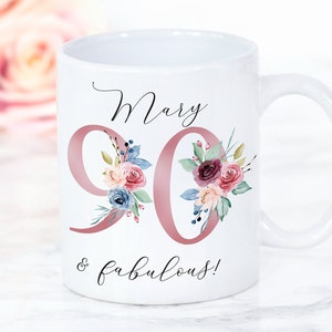Op de afbeelding: Witte keramische mok met een roze bloemenontwerp en de tekst "Mary 90 & fabulous!" erop gedrukt.